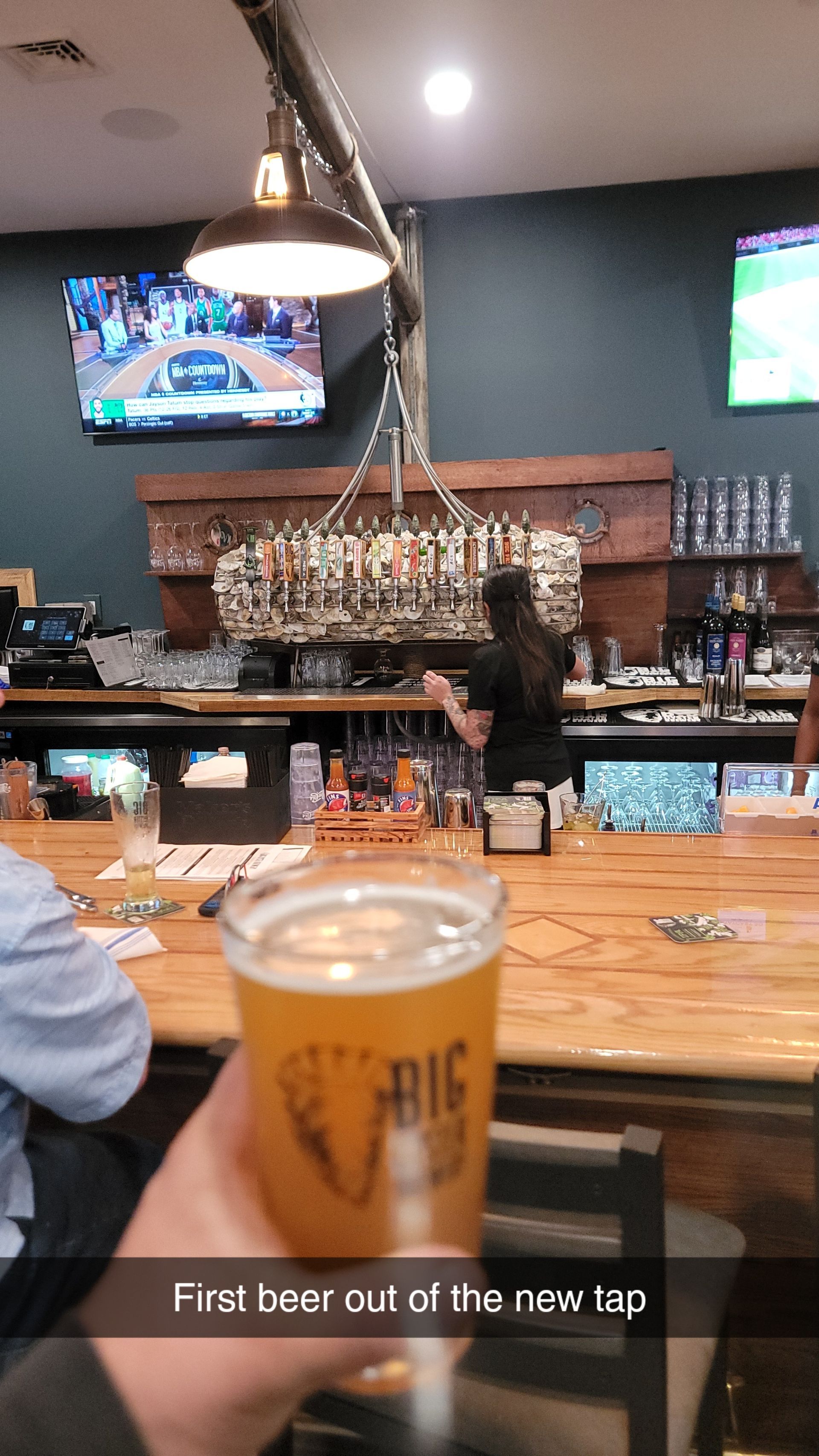 Person holding a beer at a bar; bartender behind bar.  Beer says 