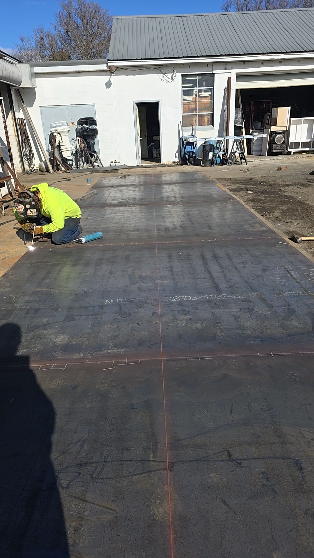 Person in yellow vest working on large metal sheets outdoors, building in background.