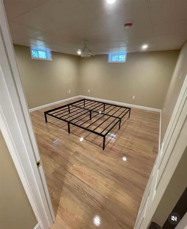 A metal bed frame centered on a light-colored wood floor in a spacious, empty basement room with two small windows.