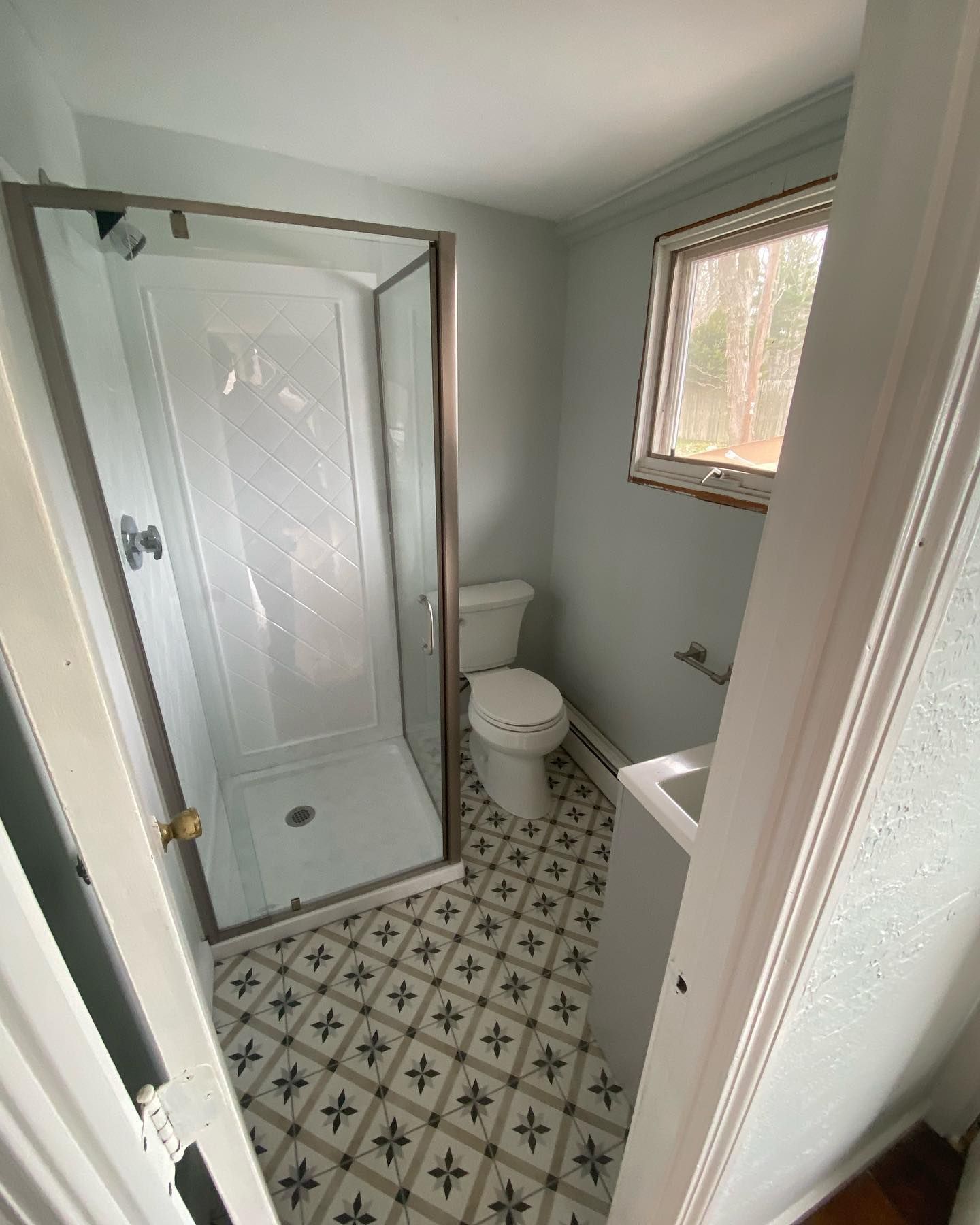 A small bathroom with light gray walls, a white shower stall, a toilet, and patterned tiled flooring.