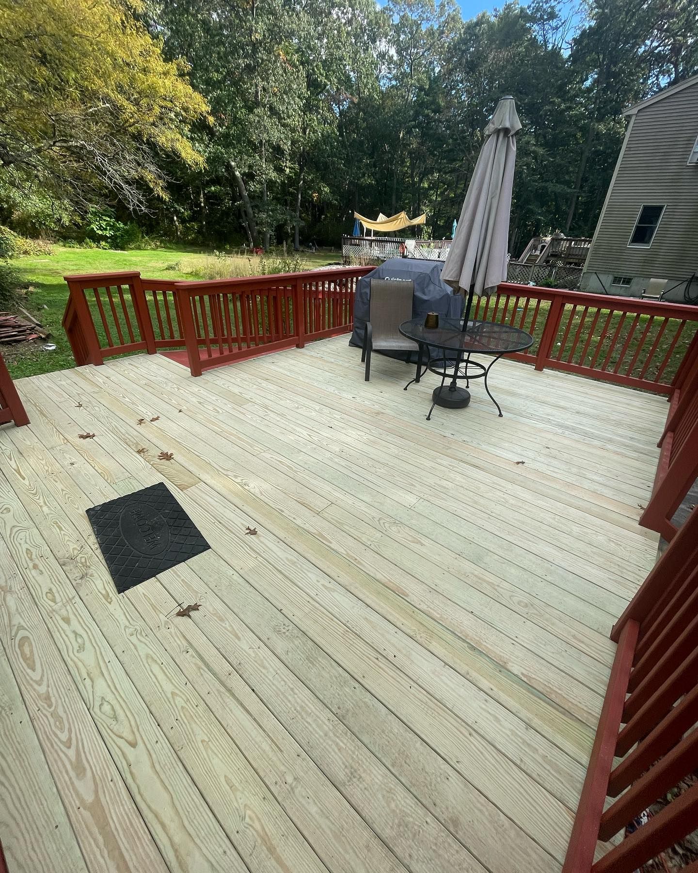 A wooden deck with red railings, a small round table with chairs, and a folded umbrella, set in a backyard.