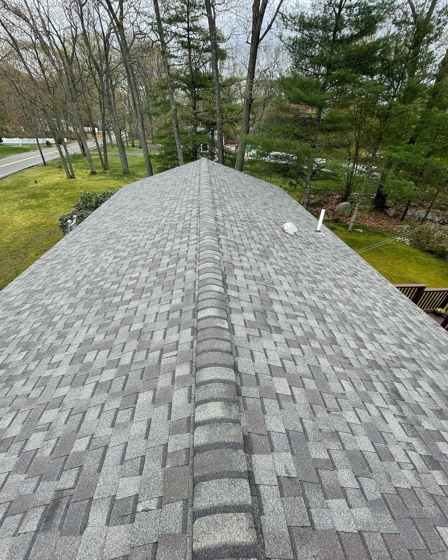 A view looking down the peaked ridge of a gray asphalt shingle roof toward a wooded yard.