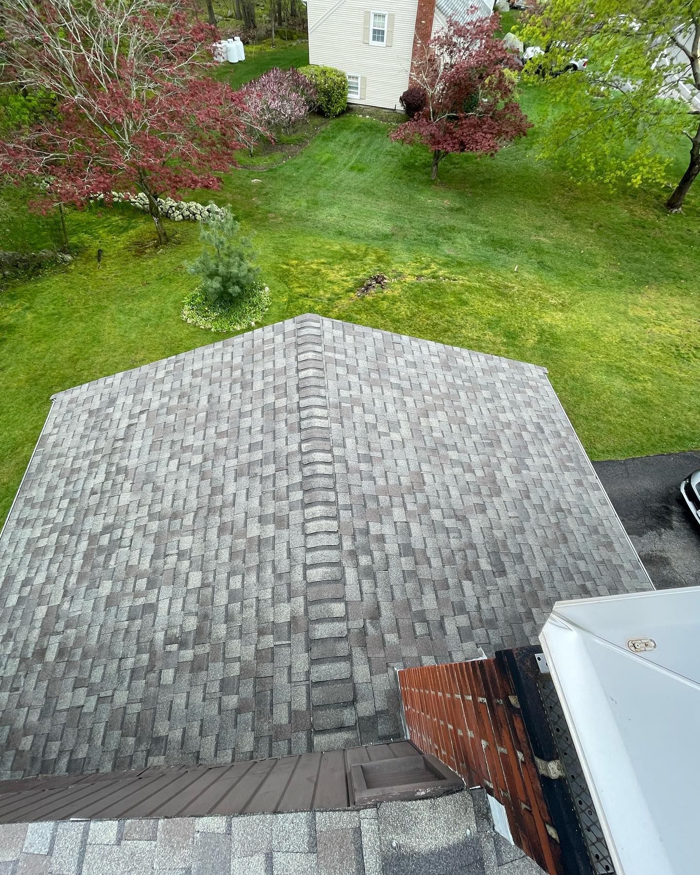 High-angle view of a grey shingled residential roof with a chimney, overlooking a green backyard with trees.