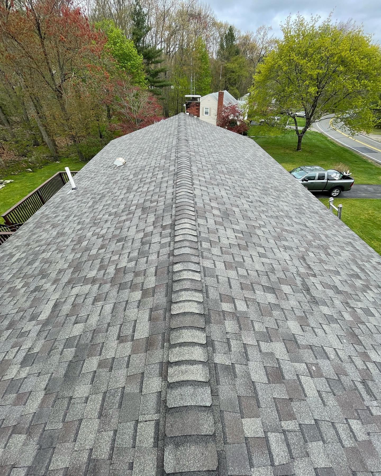 A high-angle view of a gray shingled roof with a central ridge cap, surrounded by trees and a neighborhood street.