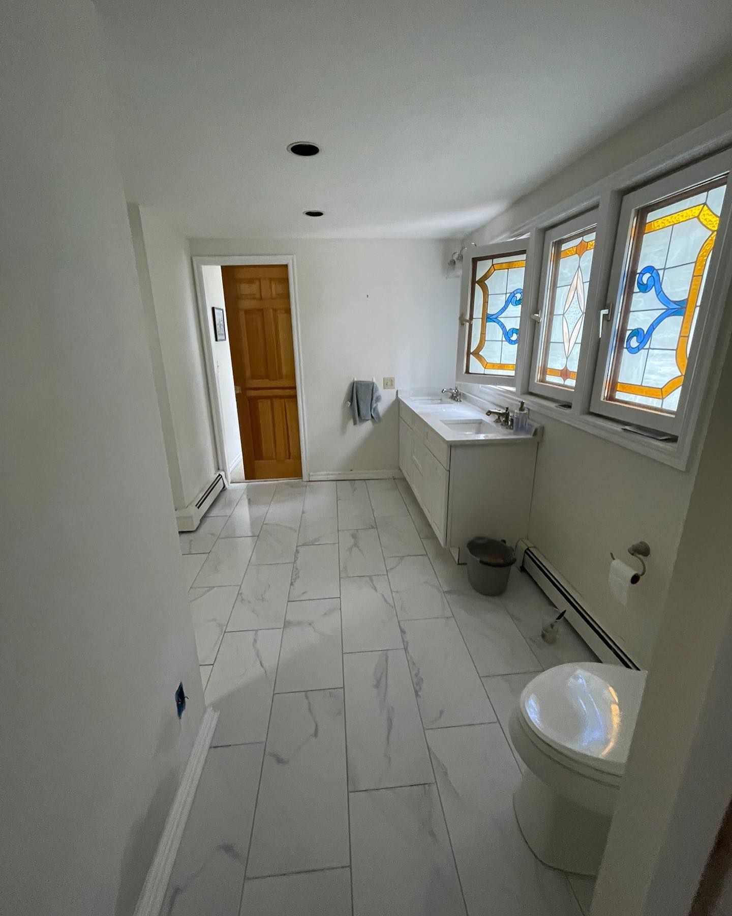 A bright bathroom with white marble-style floor tiles, a double vanity with stained glass windows, and a white toilet.
