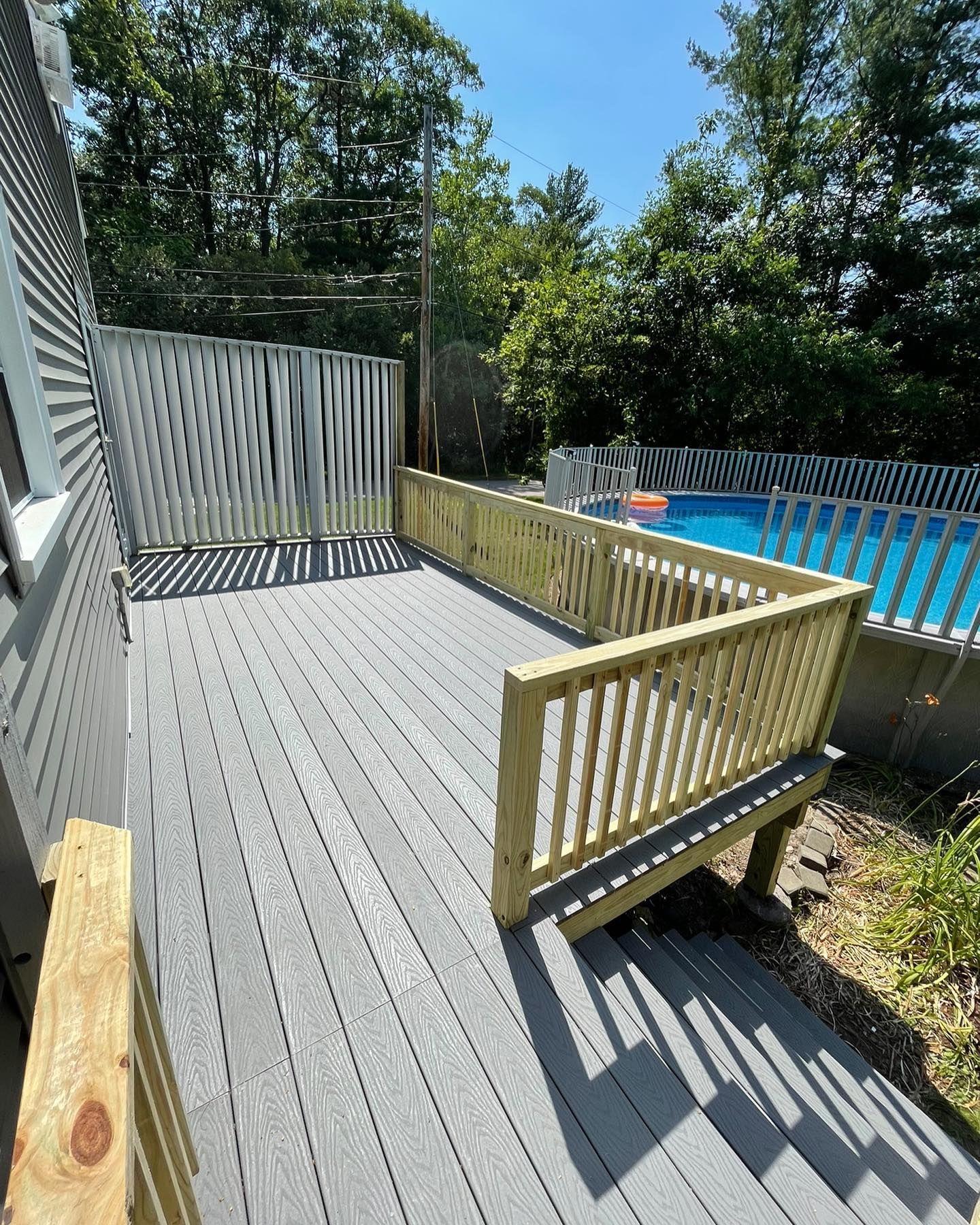 A gray composite deck with wooden railings leads to an above-ground pool in a backyard setting on a sunny day.