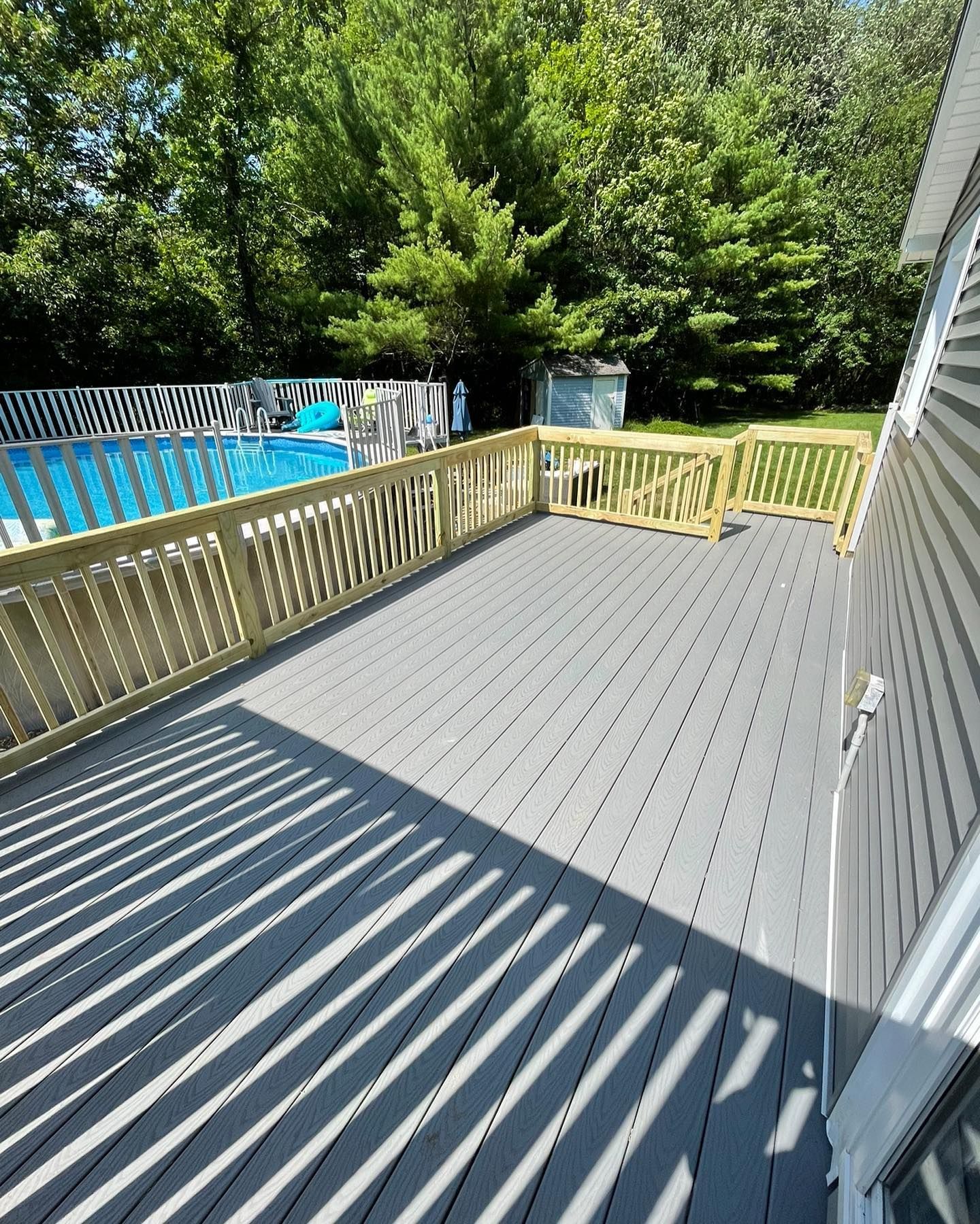 Gray composite deck with wooden railings next to a house, overlooking an above-ground swimming pool and trees.