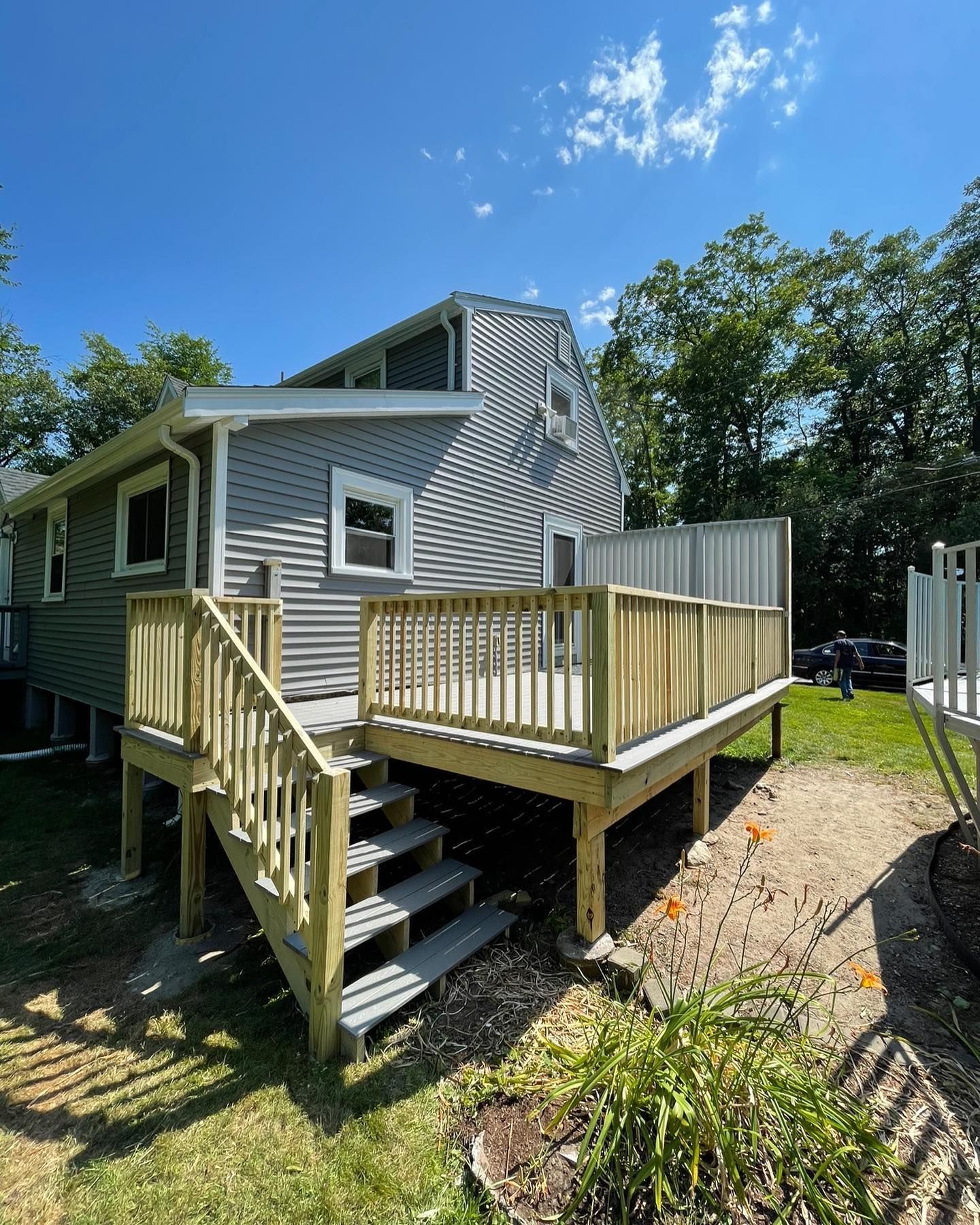 A light gray house with a new wooden deck featuring stairs and railings, set against a backdrop of green trees.