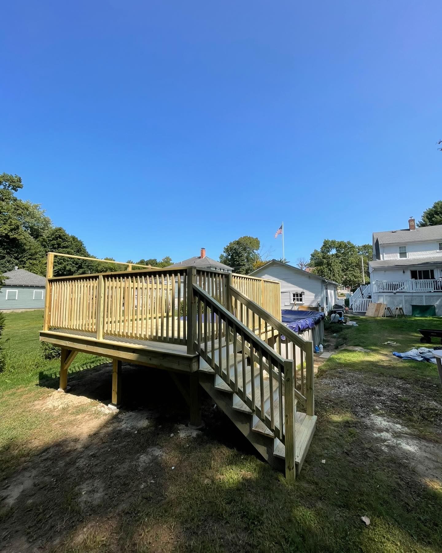 A new wooden deck with a staircase and railing stands in a sunny backyard on a clear day.