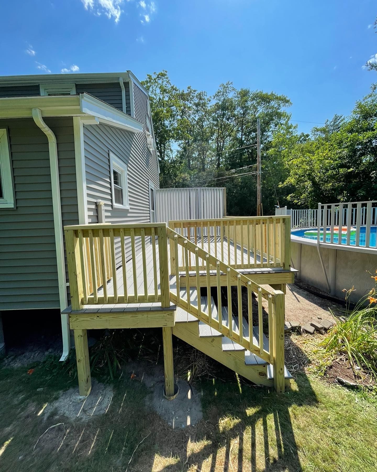 A newly built wooden deck with stairs attached to the side of a grey house, positioned near an above-ground swimming pool.