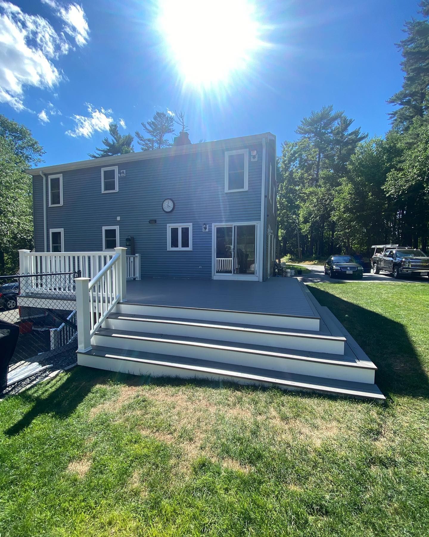 Back view of a grey two-story house with a large new grey deck and wide white stairs in a sunny, grassy backyard.