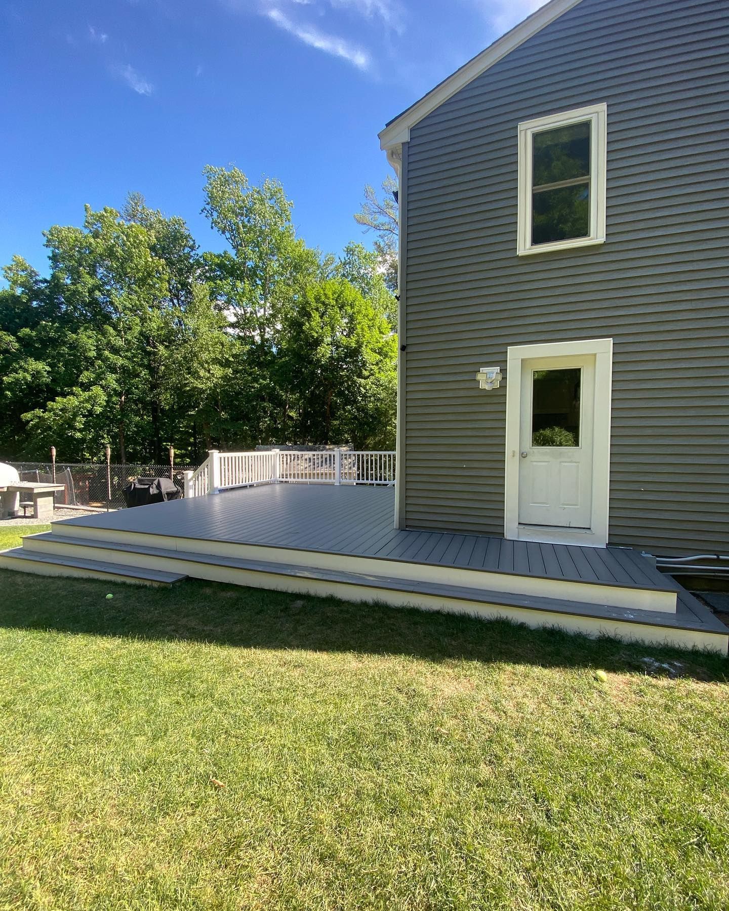 A grey-sided house with a newly installed dark grey deck and white trim, set against a backdrop of trees and a blue sky.