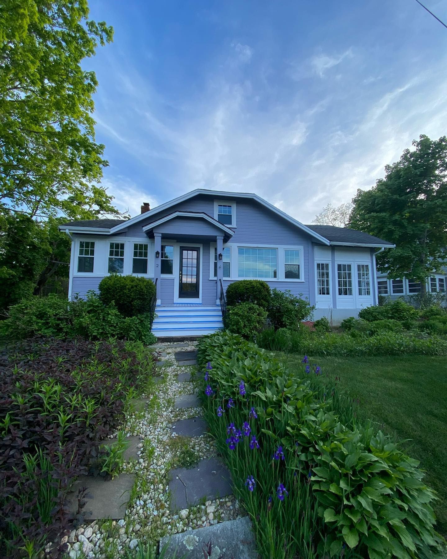 A light blue one-story house with a stone pathway, lush greenery, and blooming irises under a bright, cloudy sky.
