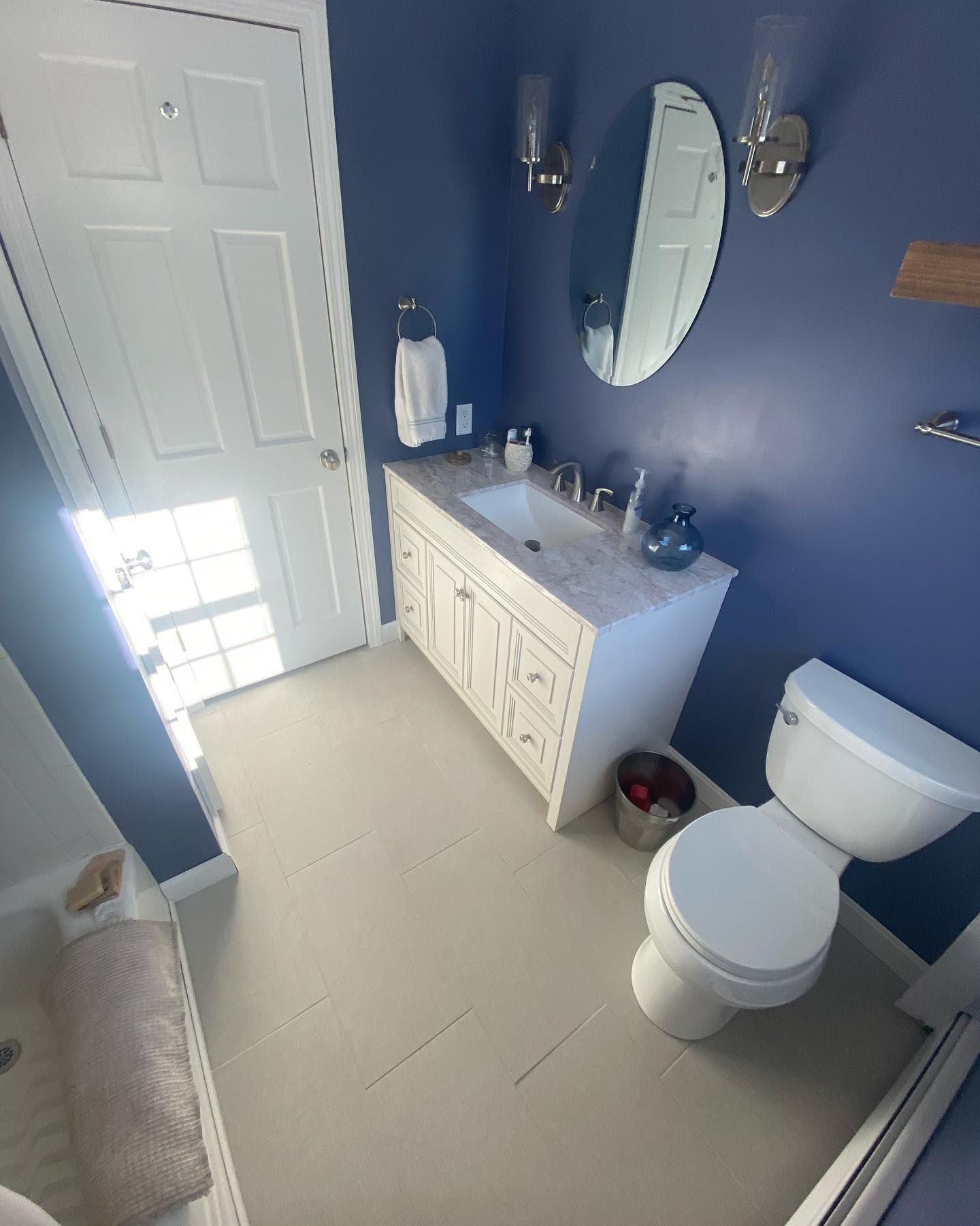 A bathroom with dark blue walls, a white vanity cabinet, an oval mirror, and a white toilet on light-colored flooring.