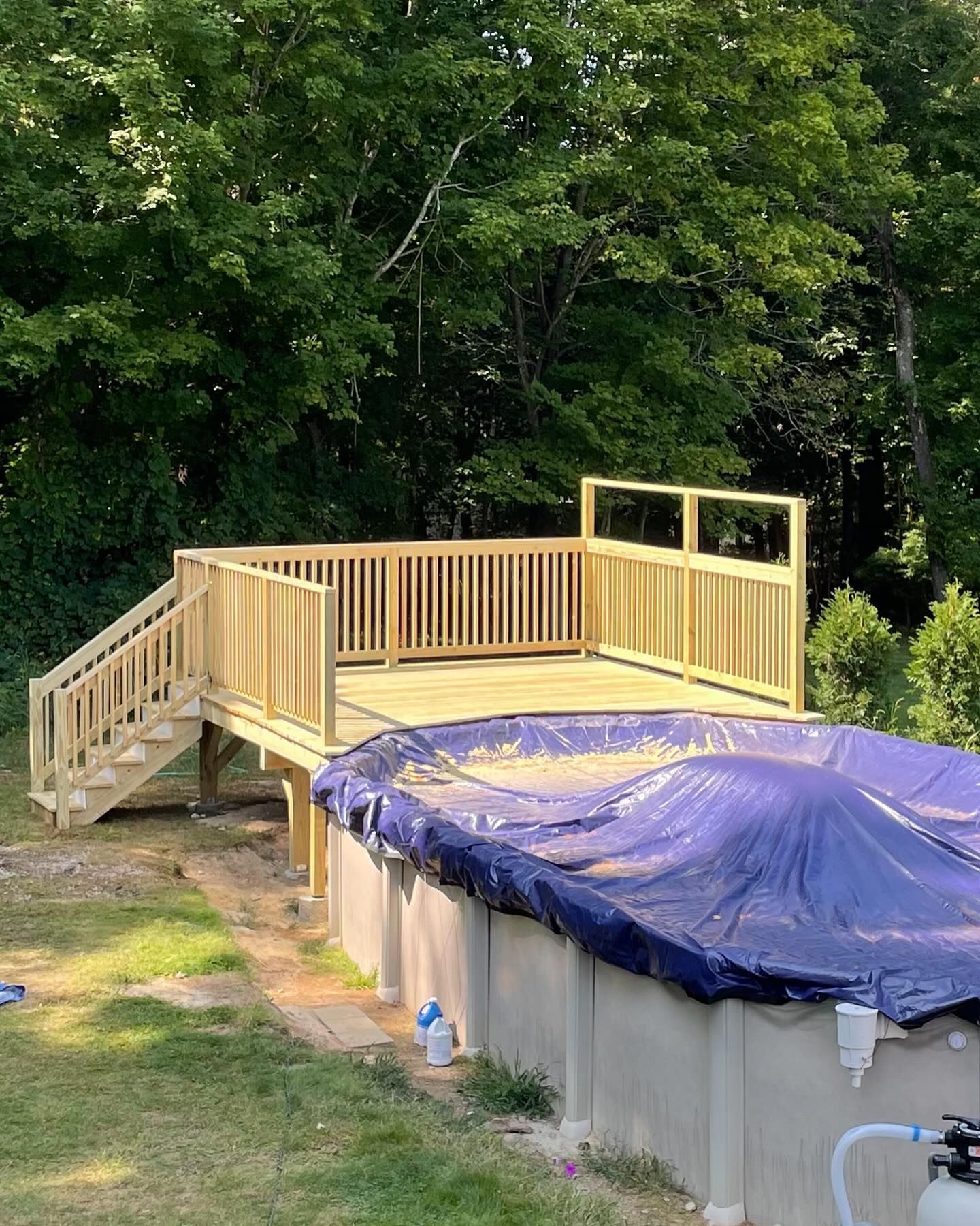 A newly built wooden deck with a staircase sits beside an above-ground swimming pool covered with a blue tarp in a yard.