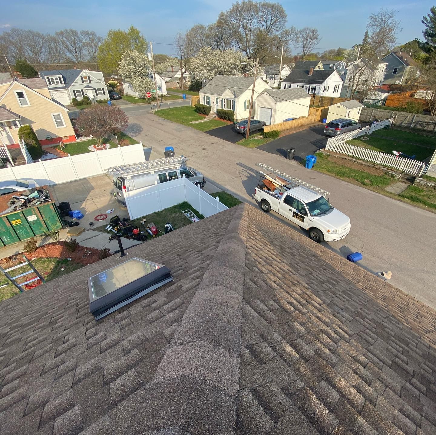 A high-angle view of a brown shingled roof under construction, with a white utility truck parked on a suburban street below.