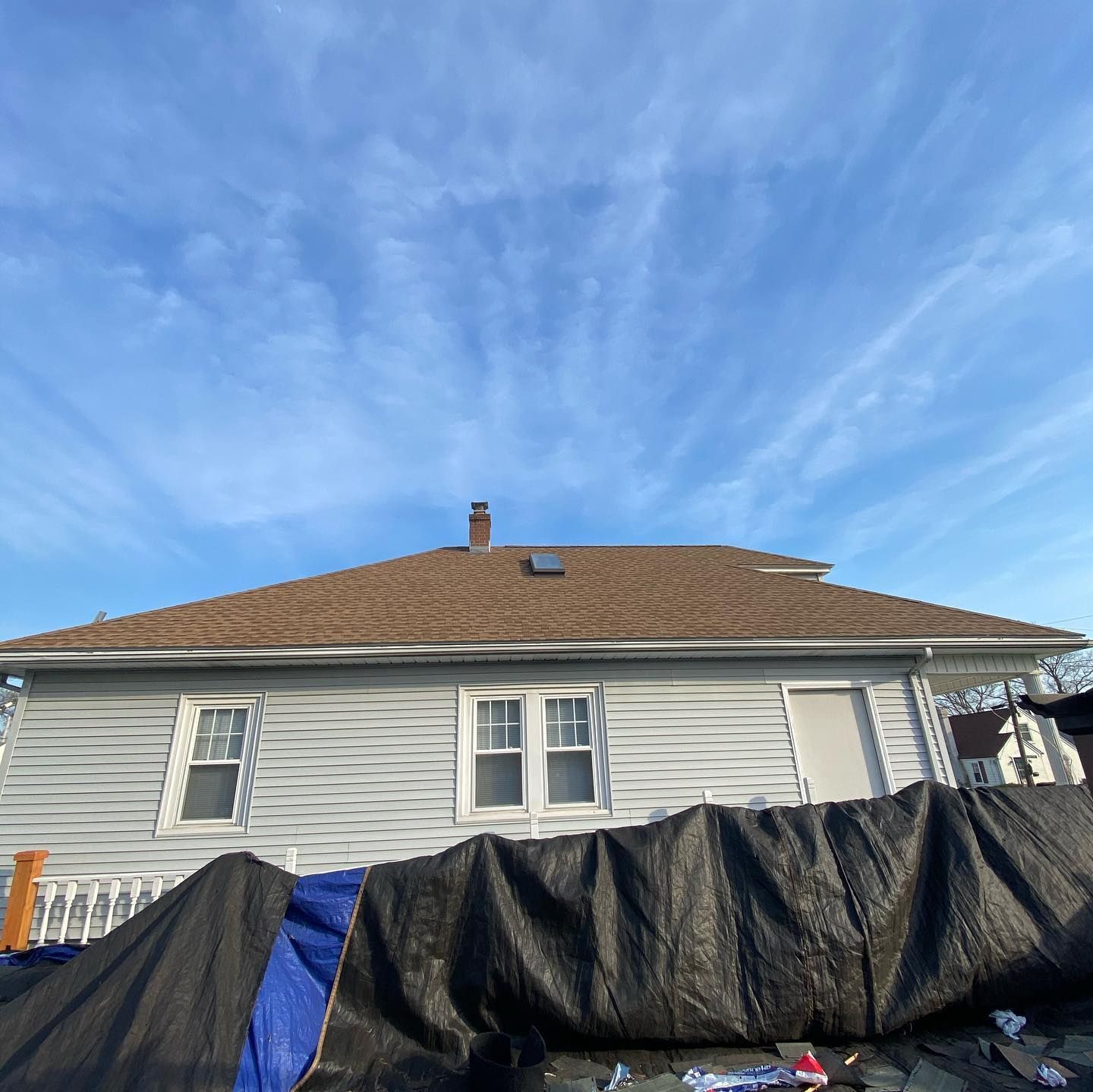 A house with wavy, light gray siding and a brown shingled roof under a blue, cloud-streaked sky, obscured by a black tarp.