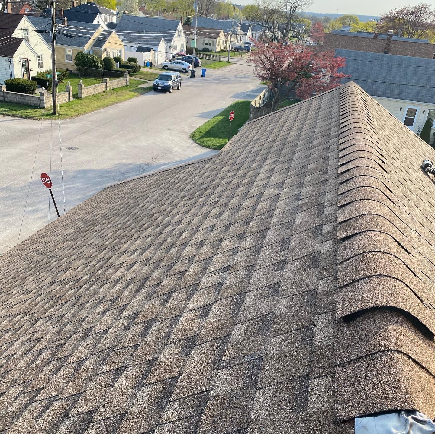 View from a residential shingled roof overlooking a suburban street with houses, cars, and trees on a sunny day.
