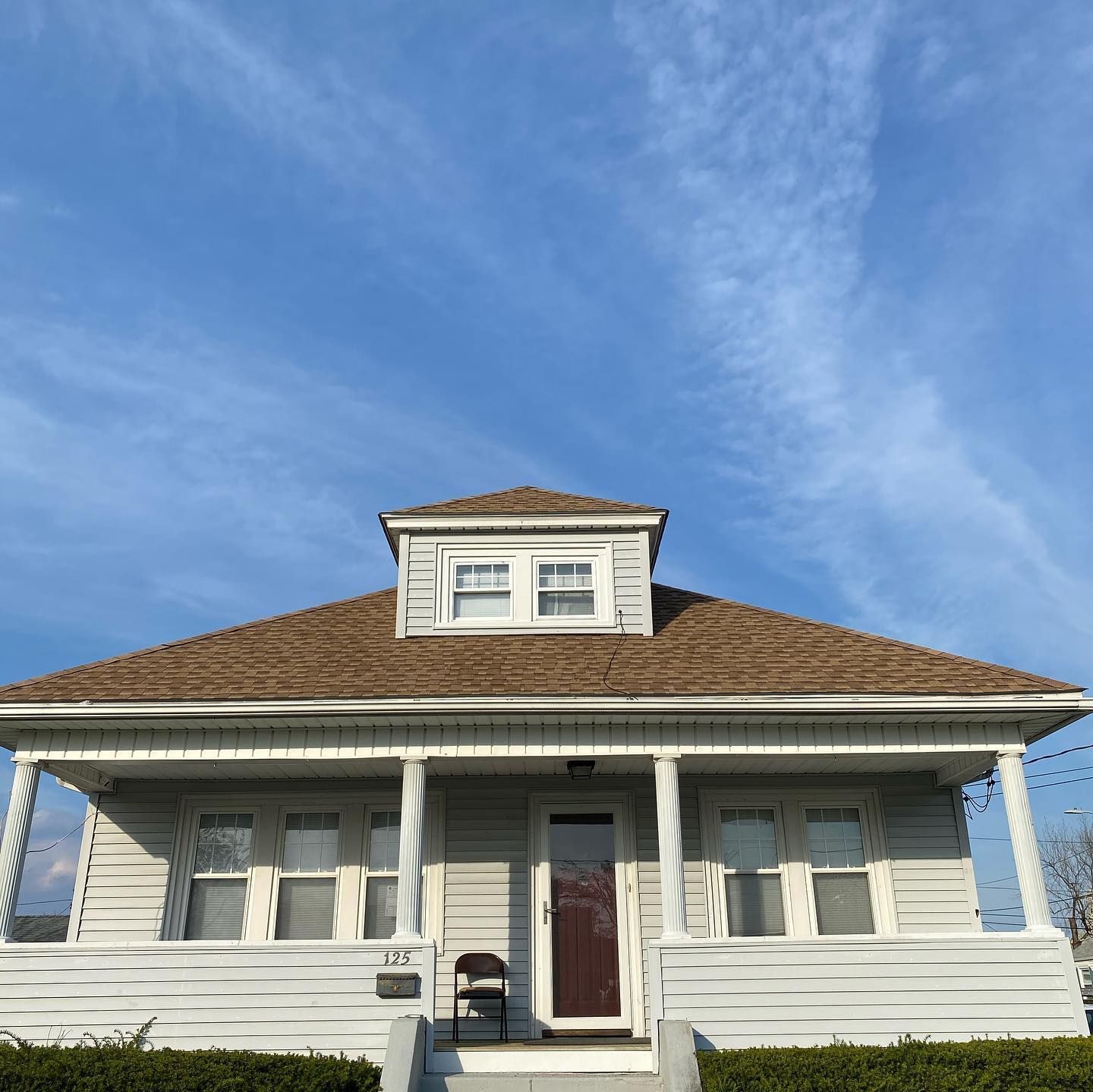 A single-story, light-colored house with a front porch, a dormer window, and a brown roof against a blue sky.