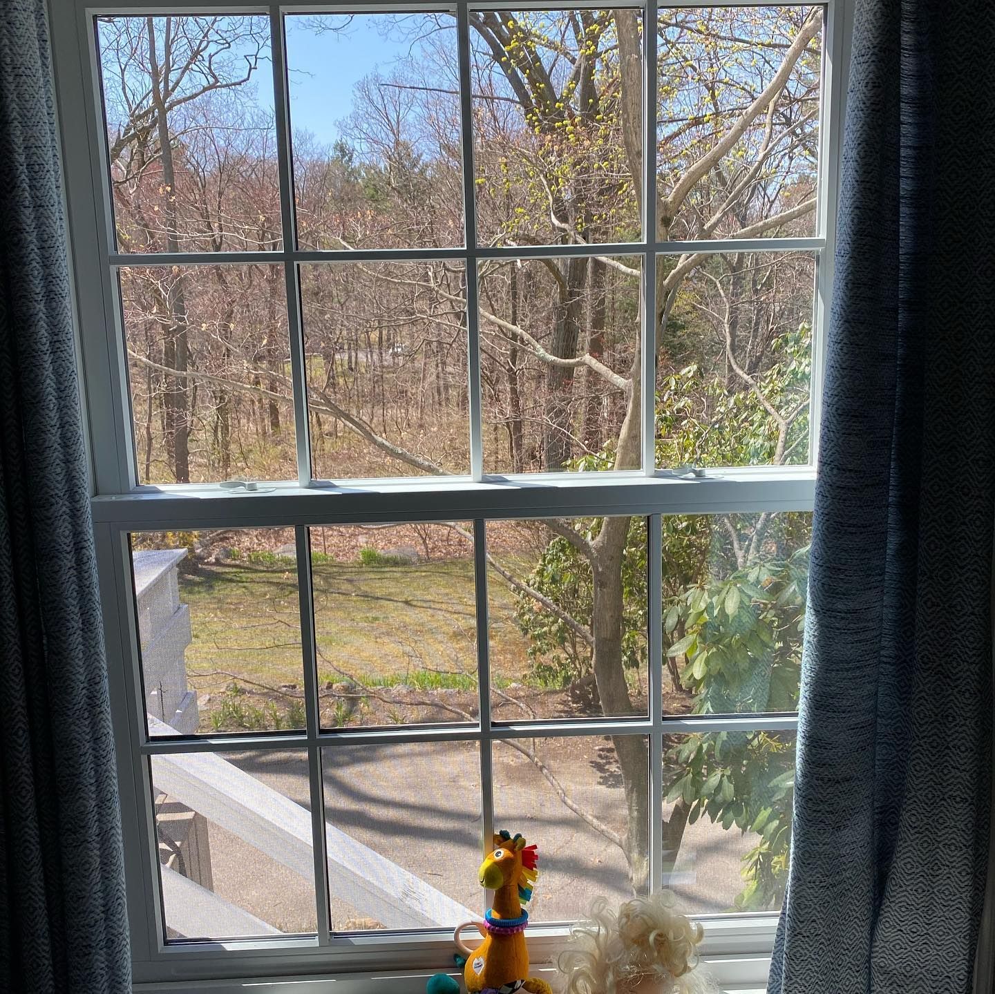 A window view showing bare trees in early spring, with a small giraffe toy and a doll sitting on the sill inside.