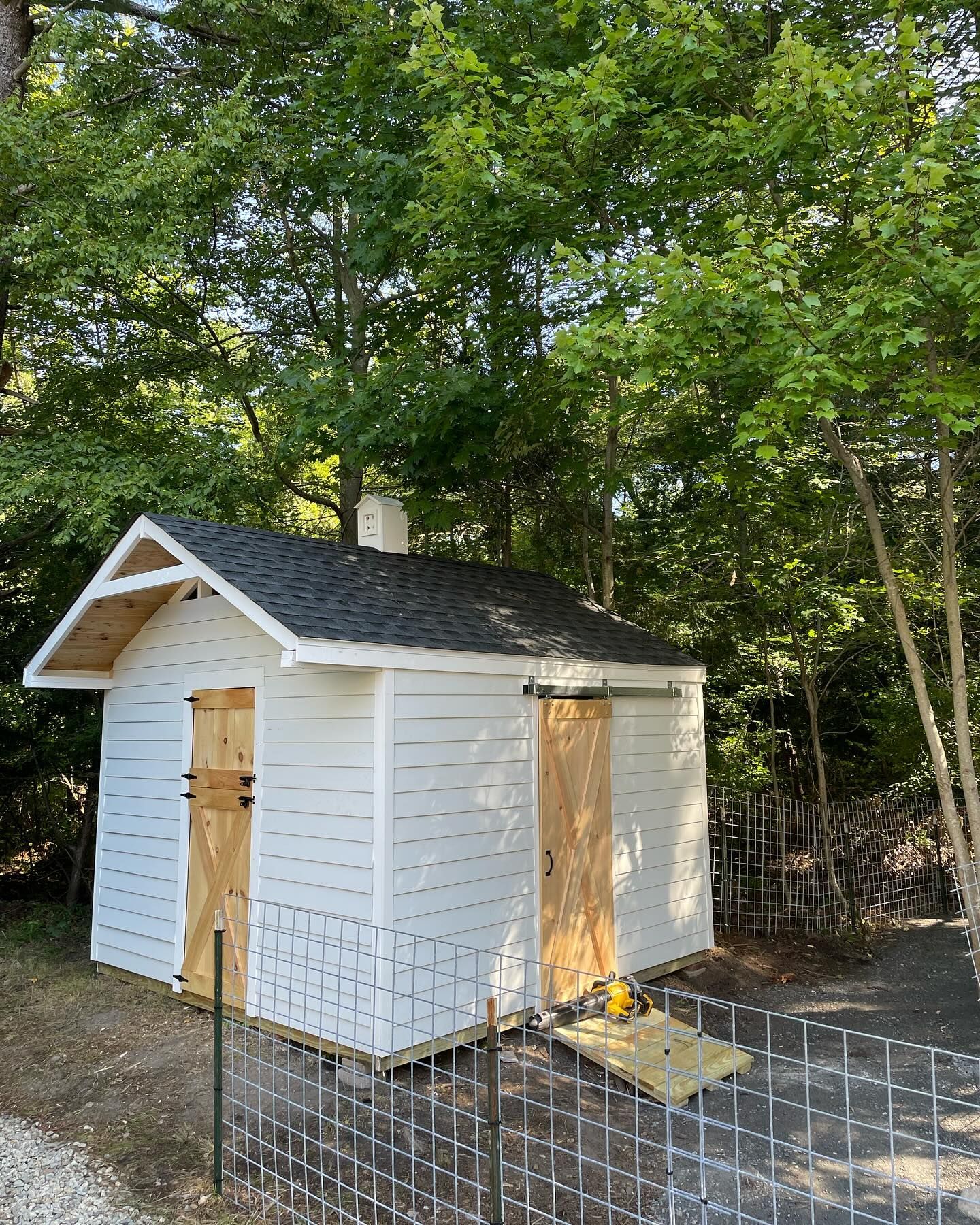 A white shed with a dark shingled roof, sliding wooden doors, and a small porch roof, set in a wooded area.