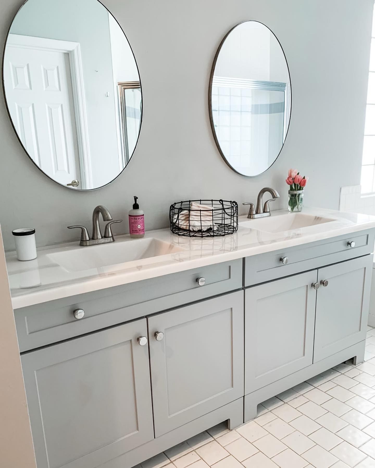 A bathroom vanity with grey cabinets, two oval mirrors, a white countertop, two sinks, and a small basket of towels.