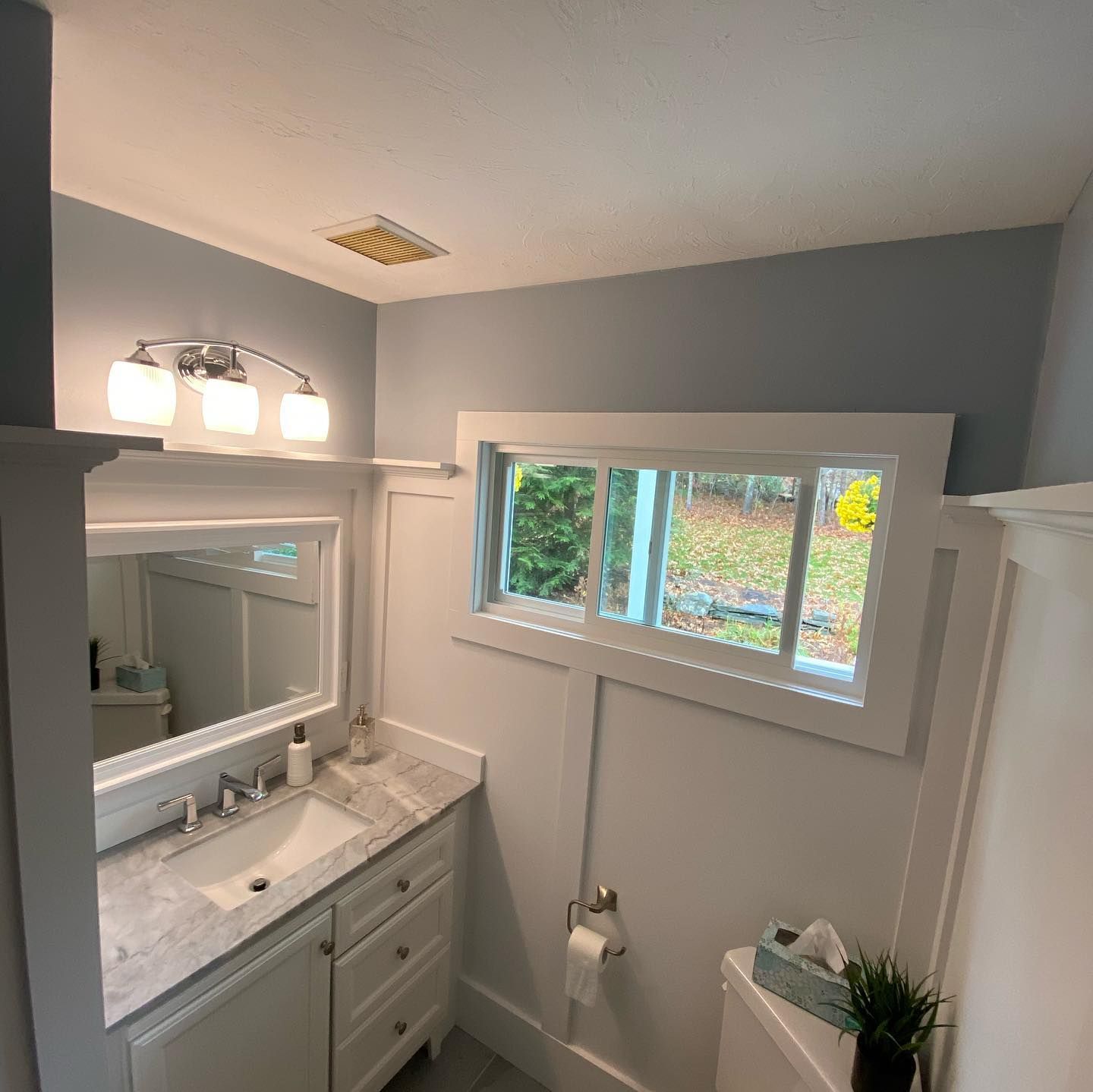 A bathroom featuring a light gray vanity with a marble countertop, an oval mirror, wall sconce lighting, and a window.