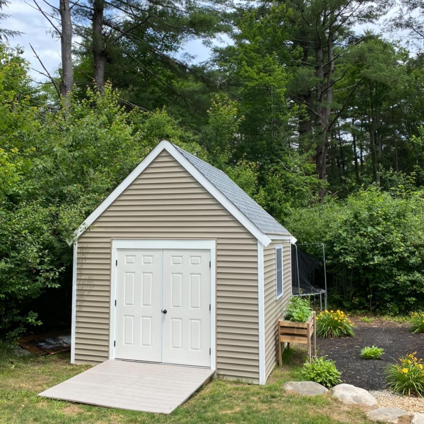 A tan shed with white trim and double doors sits on a lawn with a ramp leading up to the entrance.