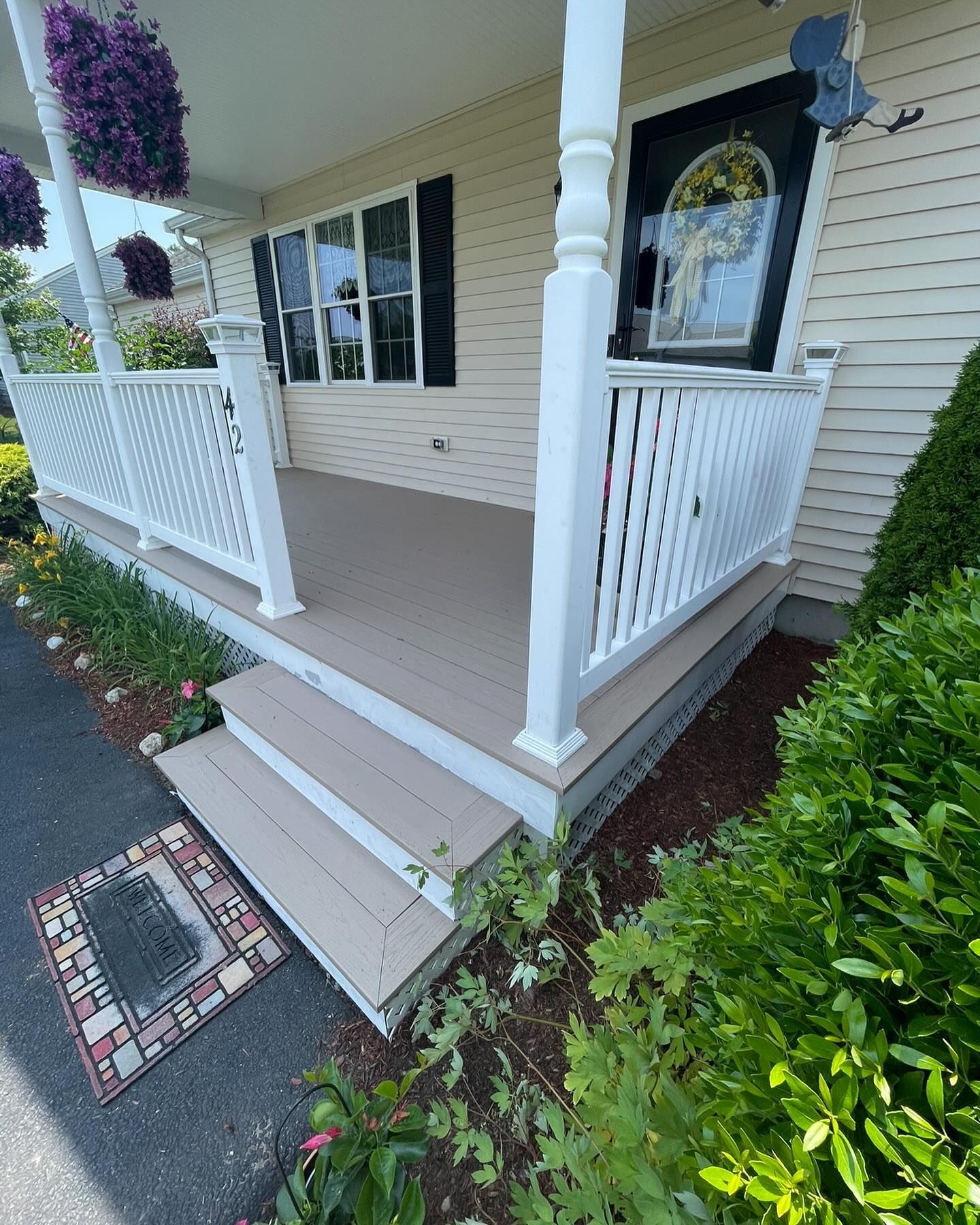 A light-colored house porch with white railings and steps, featuring hanging flowers and a door with a decorative wreath.