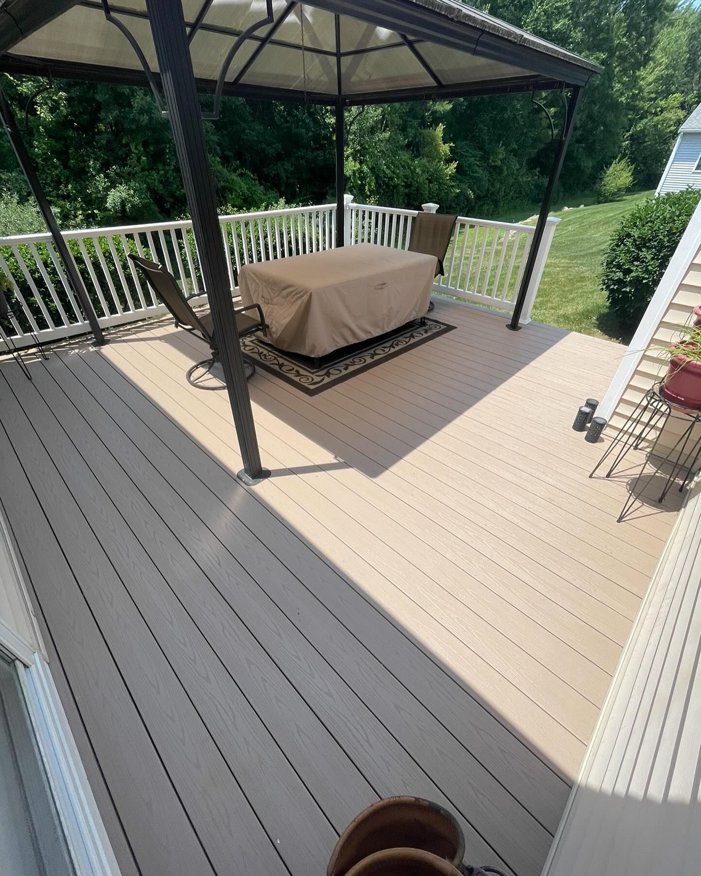 A wooden deck featuring a gazebo, a table with a tan cover, and chairs, set against a backdrop of trees and a lawn.