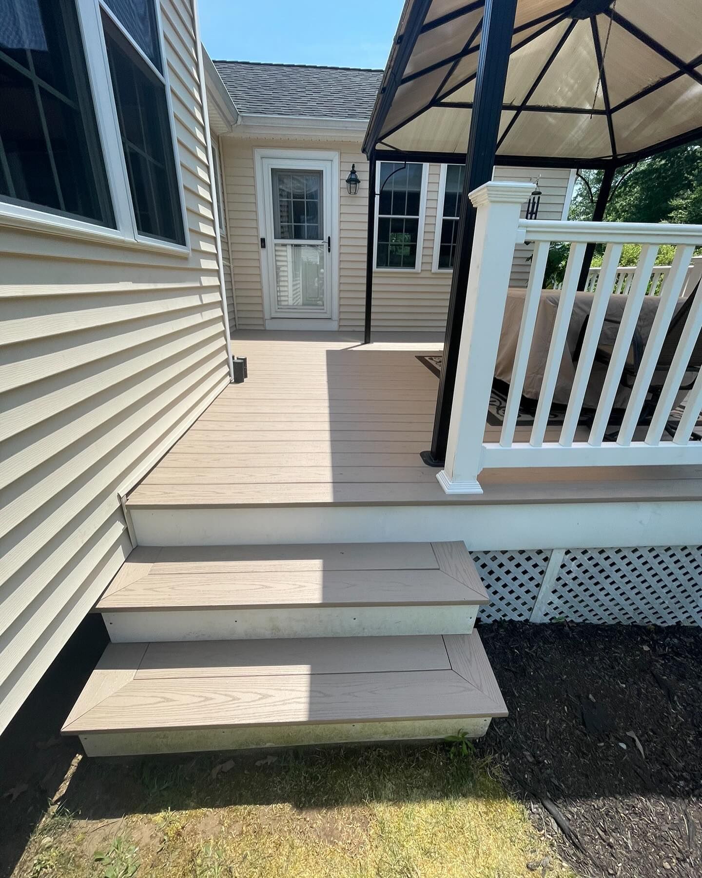 A beige house deck with two steps leading up to a white door, featuring a gazebo, white railing, and lattice skirting.