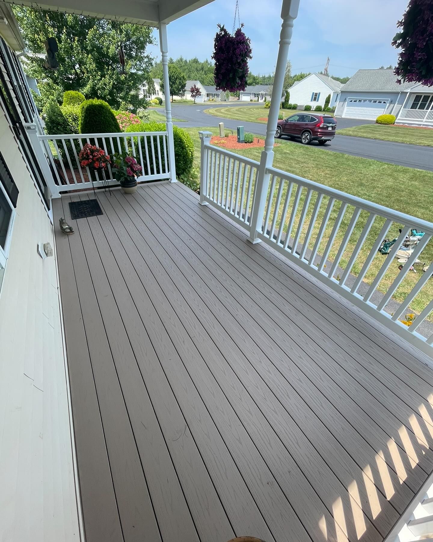 A wooden porch deck with white railings, looking out toward a suburban street with a parked car and a neighboring house.