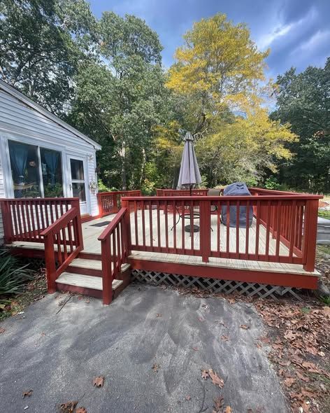 A wooden deck with red railings and stairs, featuring an umbrella and grill, sits beside a house near trees in autumn.