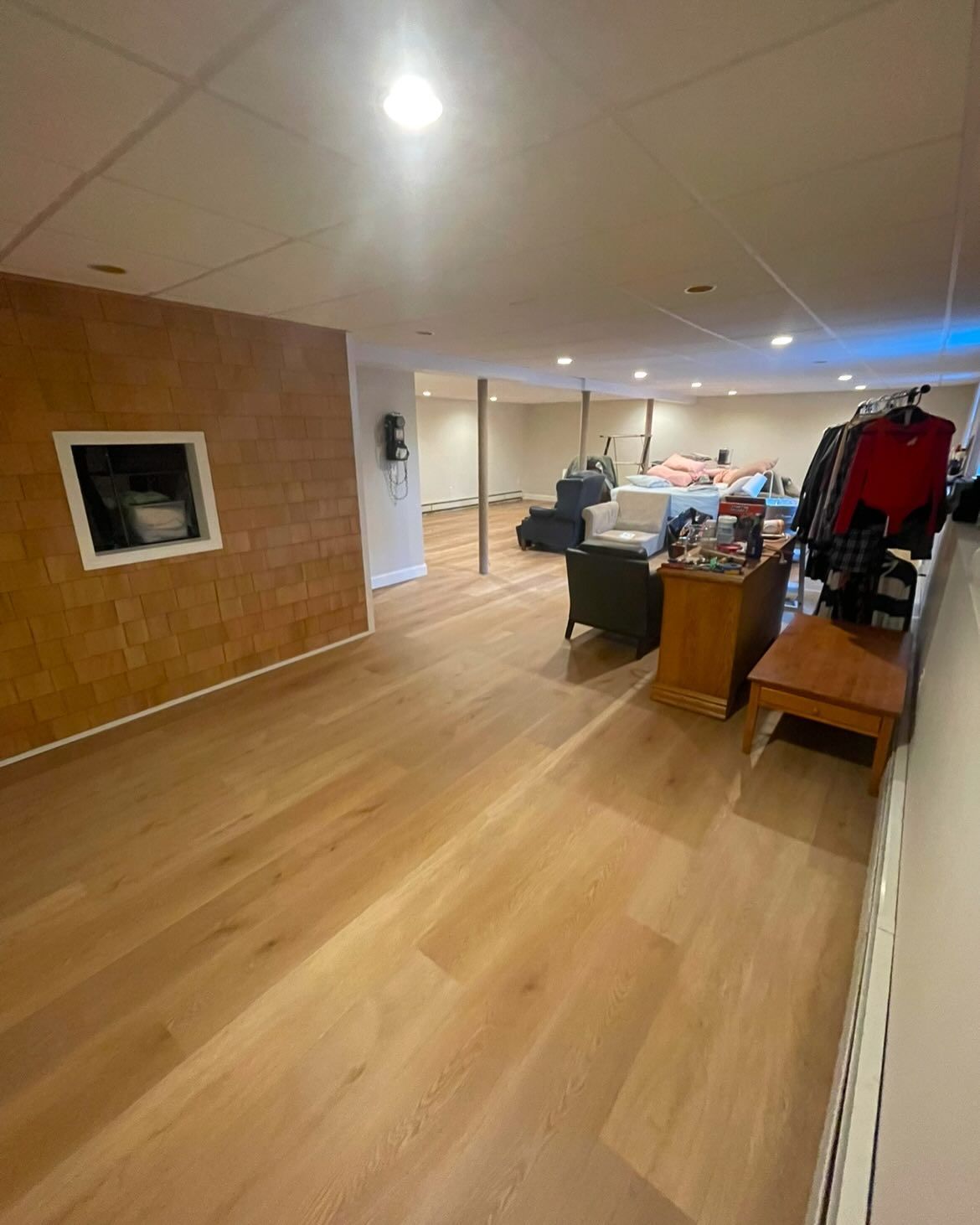 A wide-angle view of a finished basement with light wood flooring, a tan accent wall, and furniture in the background.