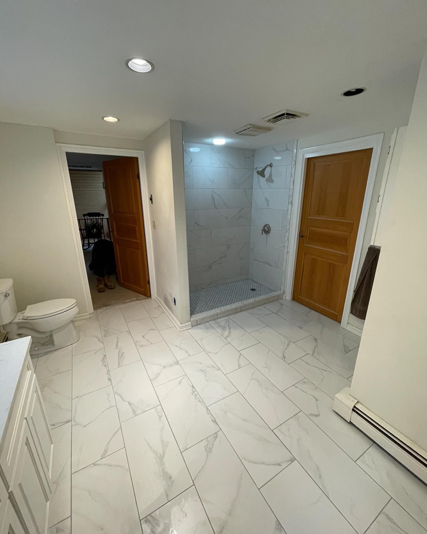 A bright bathroom with white marble-style tiled floors, a walk-in glass shower, a toilet, and two wooden doors.