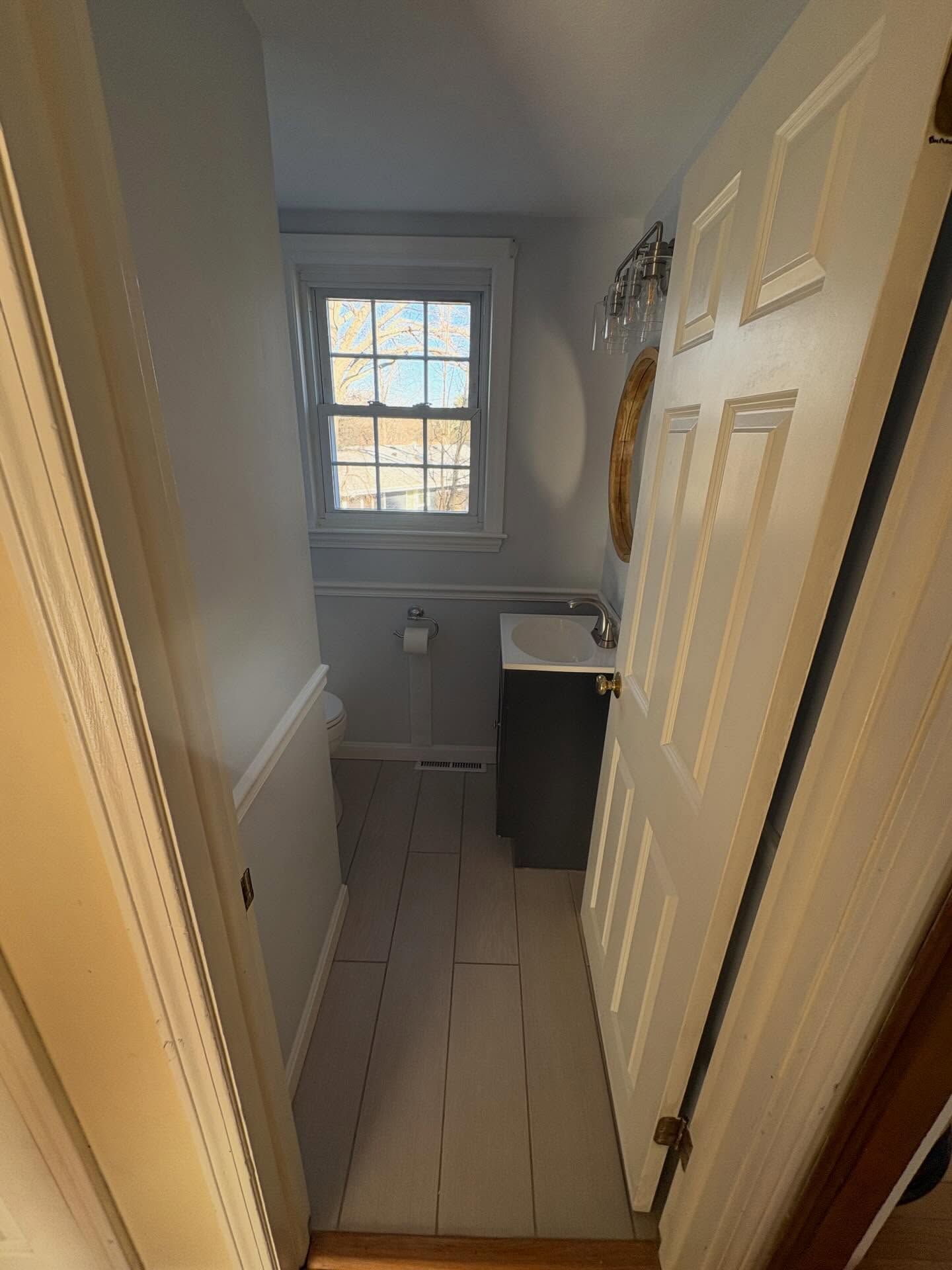 A doorway opens to a small bathroom featuring gray walls, painted wood floors, a dark vanity, and a window.