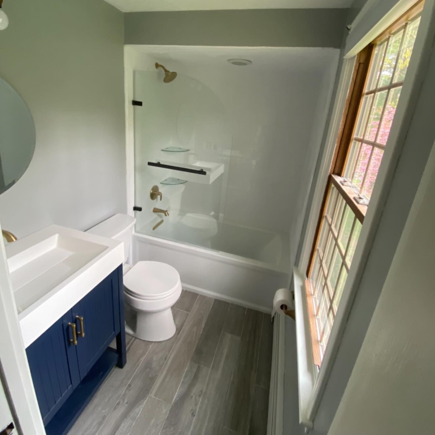 A small, modern bathroom featuring a blue vanity, a toilet, and a white bathtub-shower combo with glass shelving.