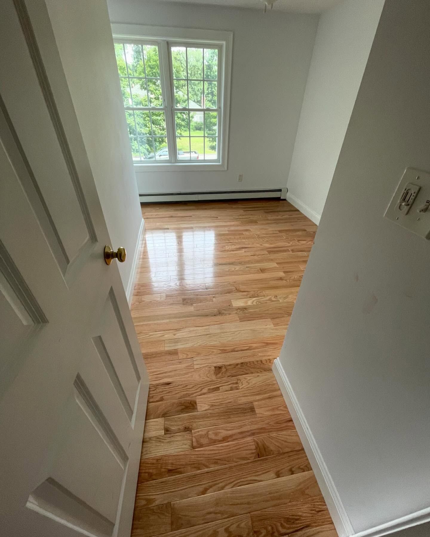 A room view from a doorway, featuring polished hardwood floors, white walls, and a large window.