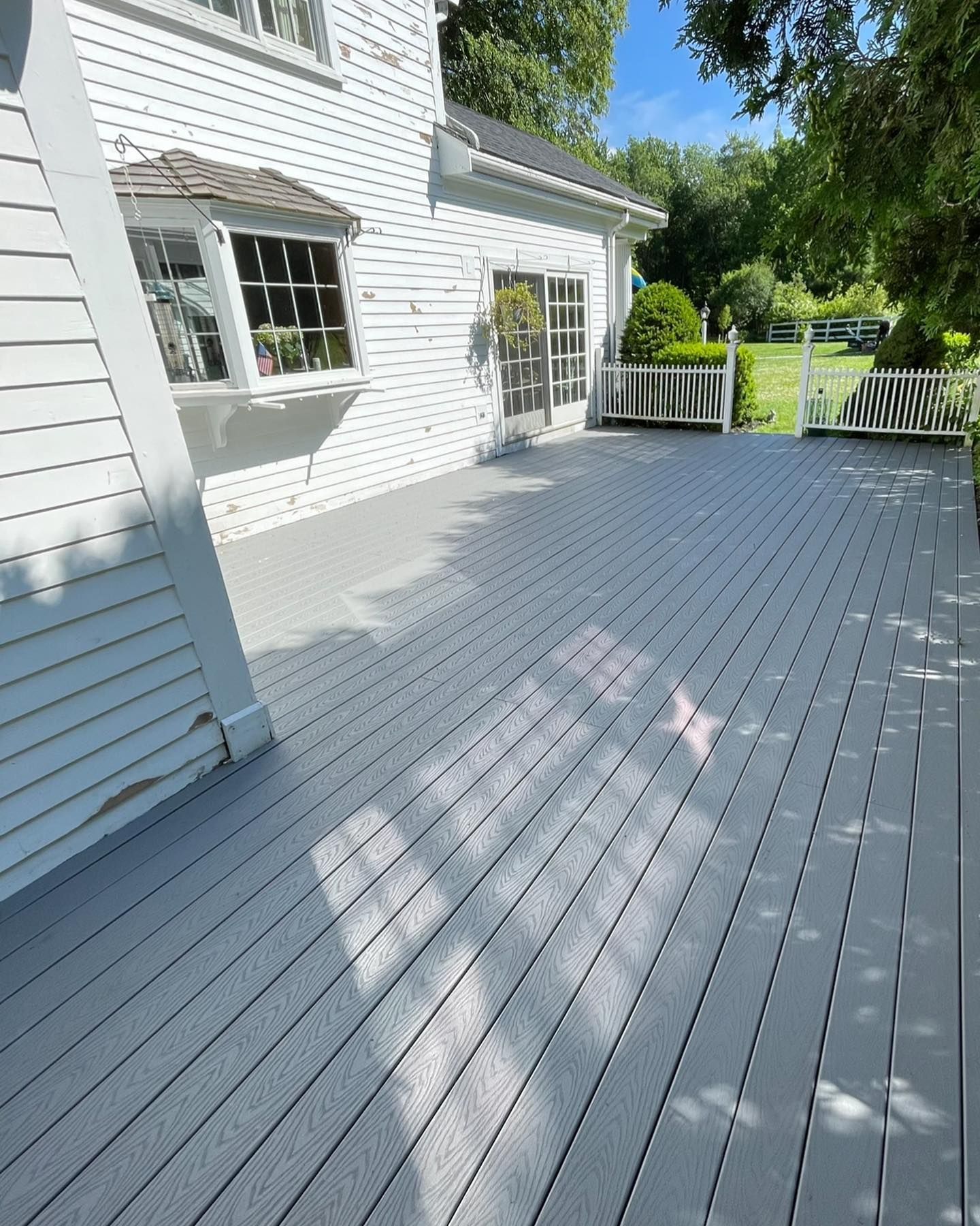 A freshly stained light-gray wooden deck attached to the side of a white house with a bay window and sliding glass door.