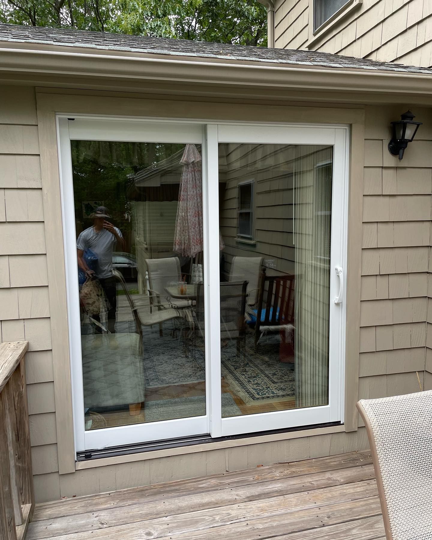White sliding glass door on a shingled house exterior, reflecting a patio area with furniture and a person standing inside.
