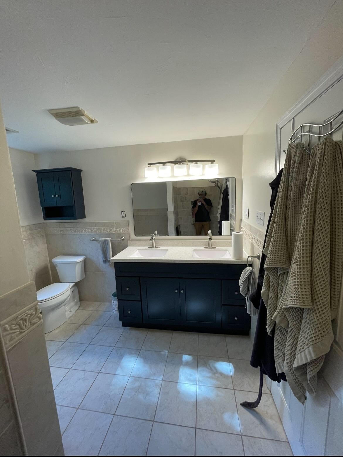 A bathroom featuring a dark blue vanity with a double sink, a matching wall cabinet, a toilet, and towels hanging on wall.