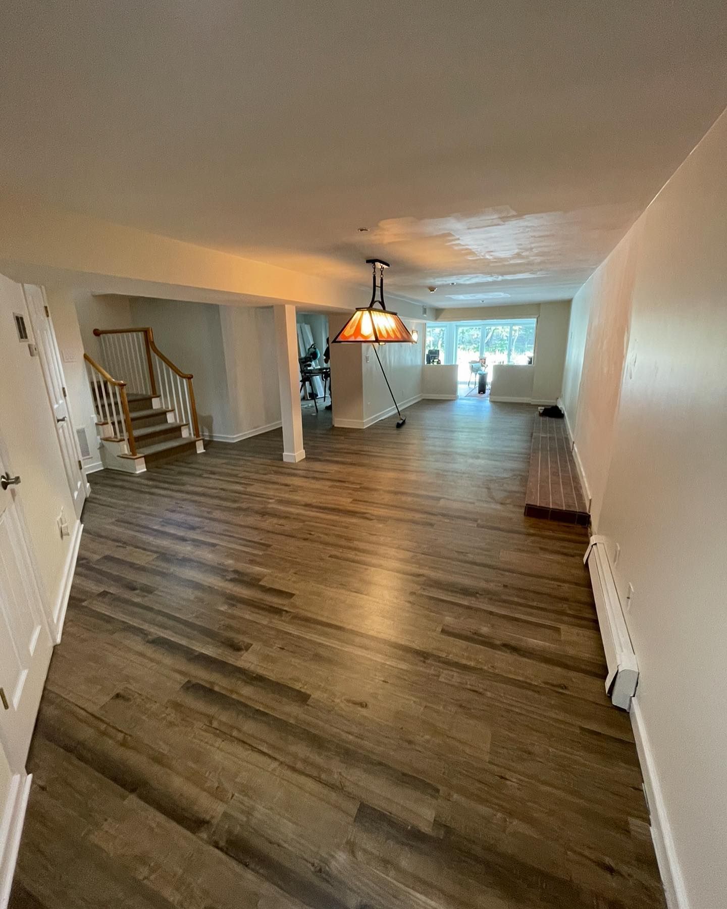 A long, empty basement room featuring wood-style flooring, white walls, a decorative hanging lamp, and a wooden staircase.
