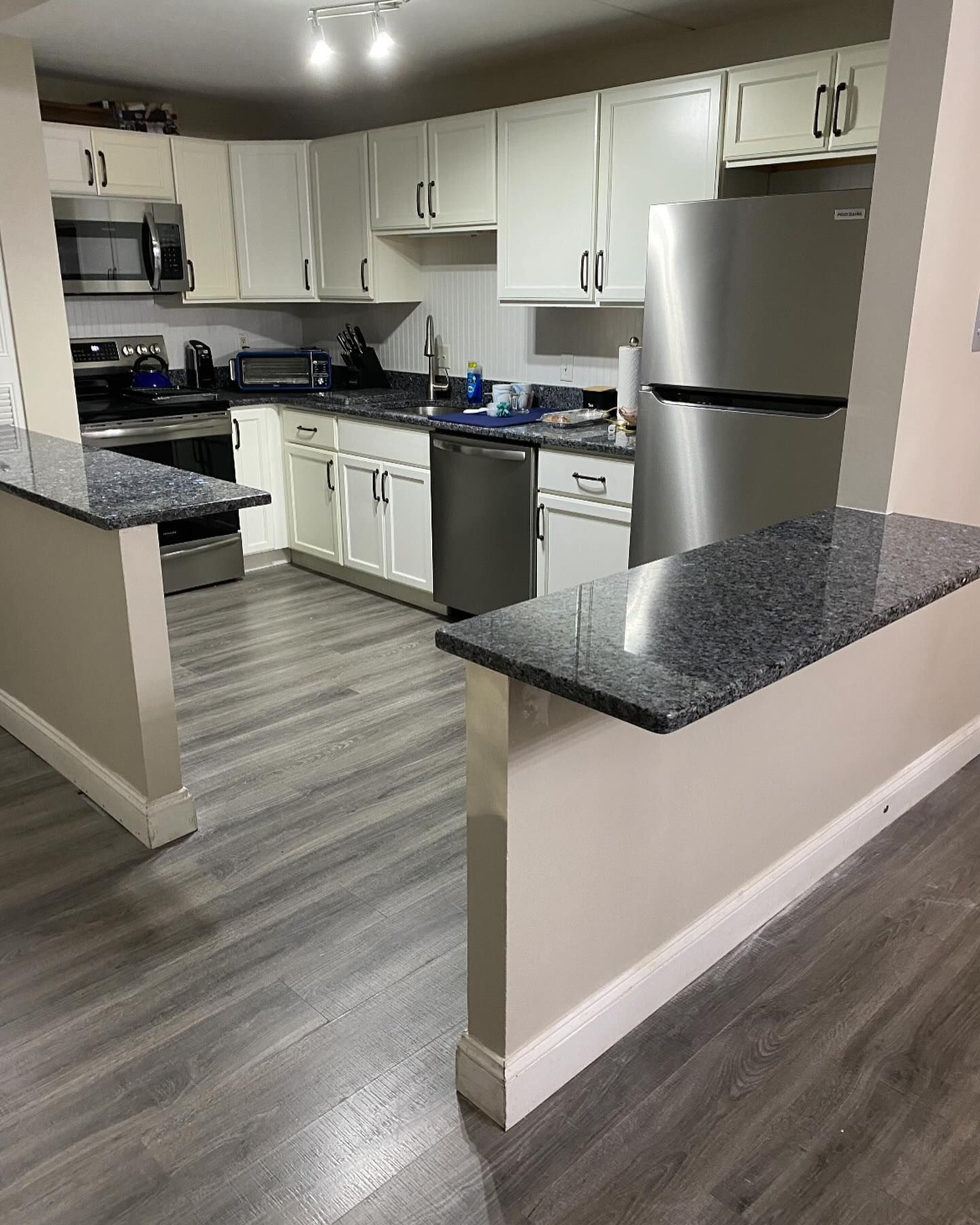 A kitchen with white cabinets, dark granite countertops, stainless steel appliances, and light gray wood-look flooring.