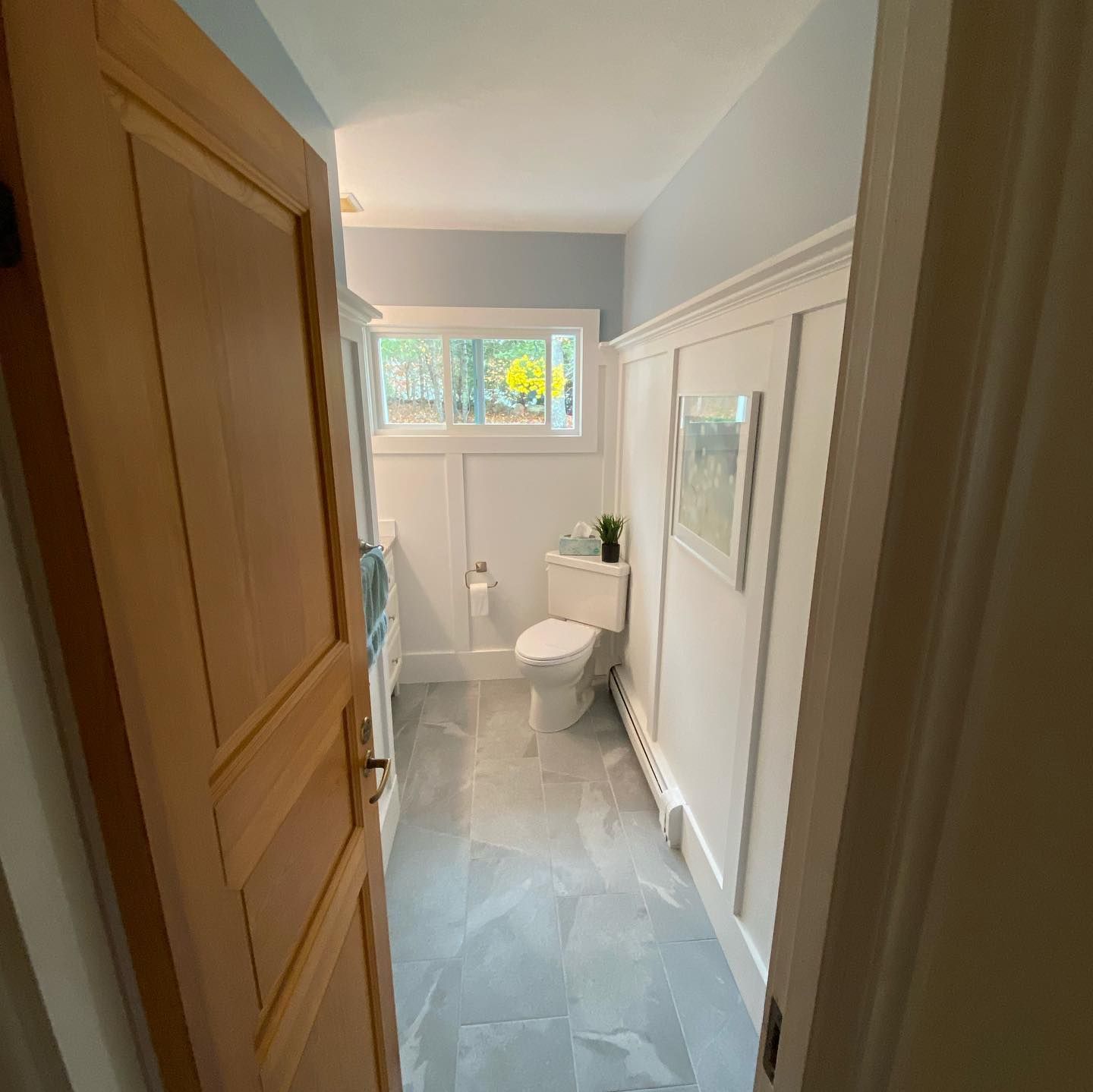 A view through an open wooden door into a bright bathroom with grey tiled floors, white wainscoting, and a white toilet.