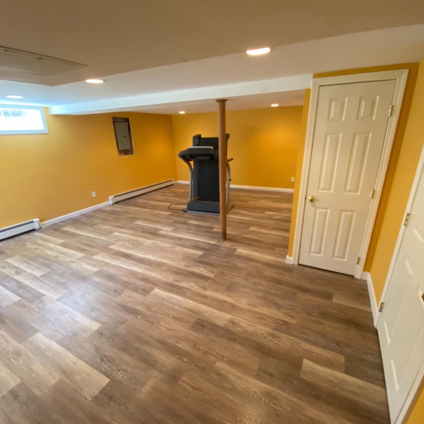 Basement with wood-look flooring, mustard-yellow walls, a treadmill near a support pole, and white doors.