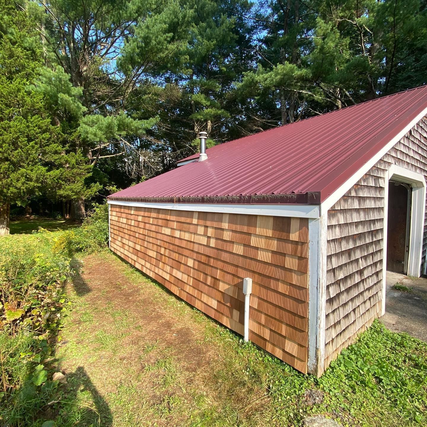 A small structure with cedar shake siding and a dark red metal roof, surrounded by green trees and foliage.