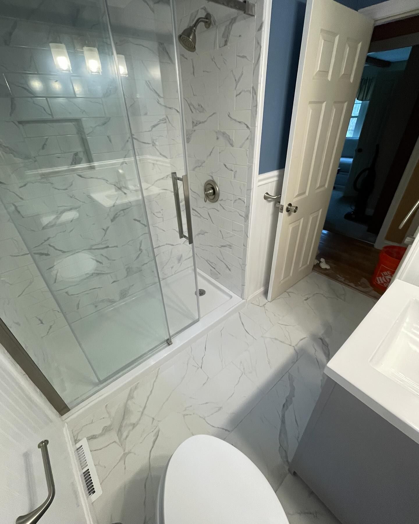 A high-angle view of a bathroom featuring a glass shower stall, marble-patterned tiled floor, white vanity, and toilet.