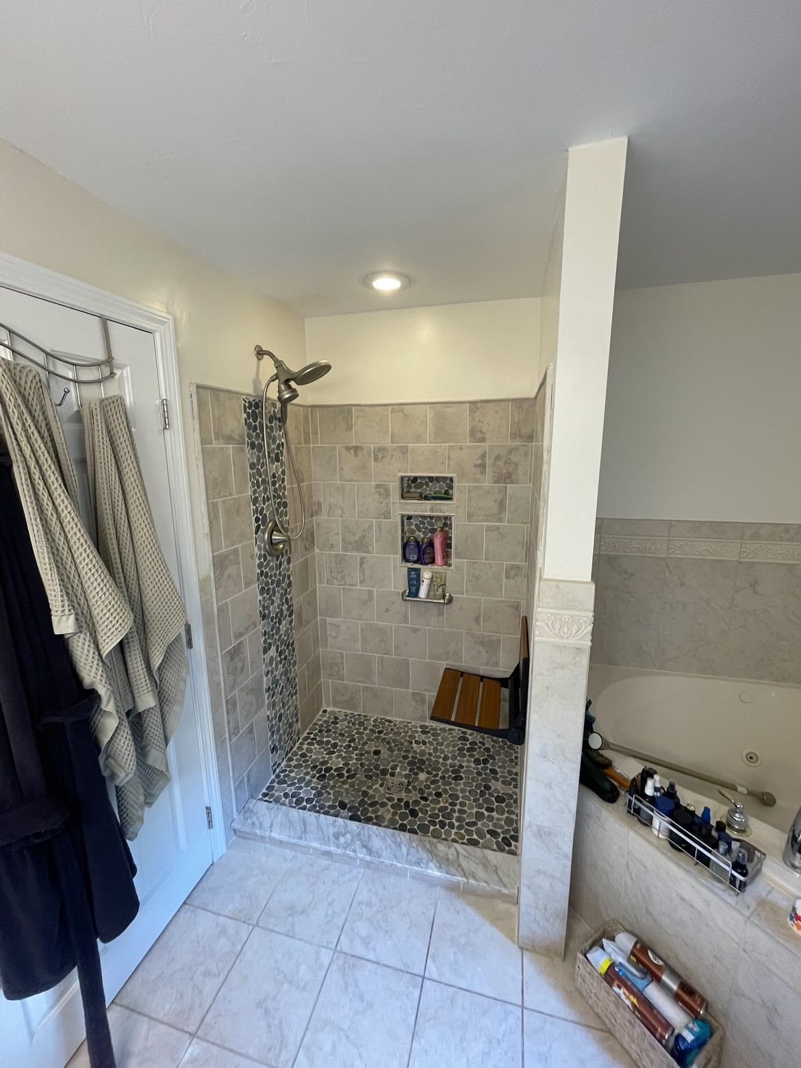 A walk-in shower with a pebble floor, stone tile walls, a built-in wooden seat, and a metal shower head.