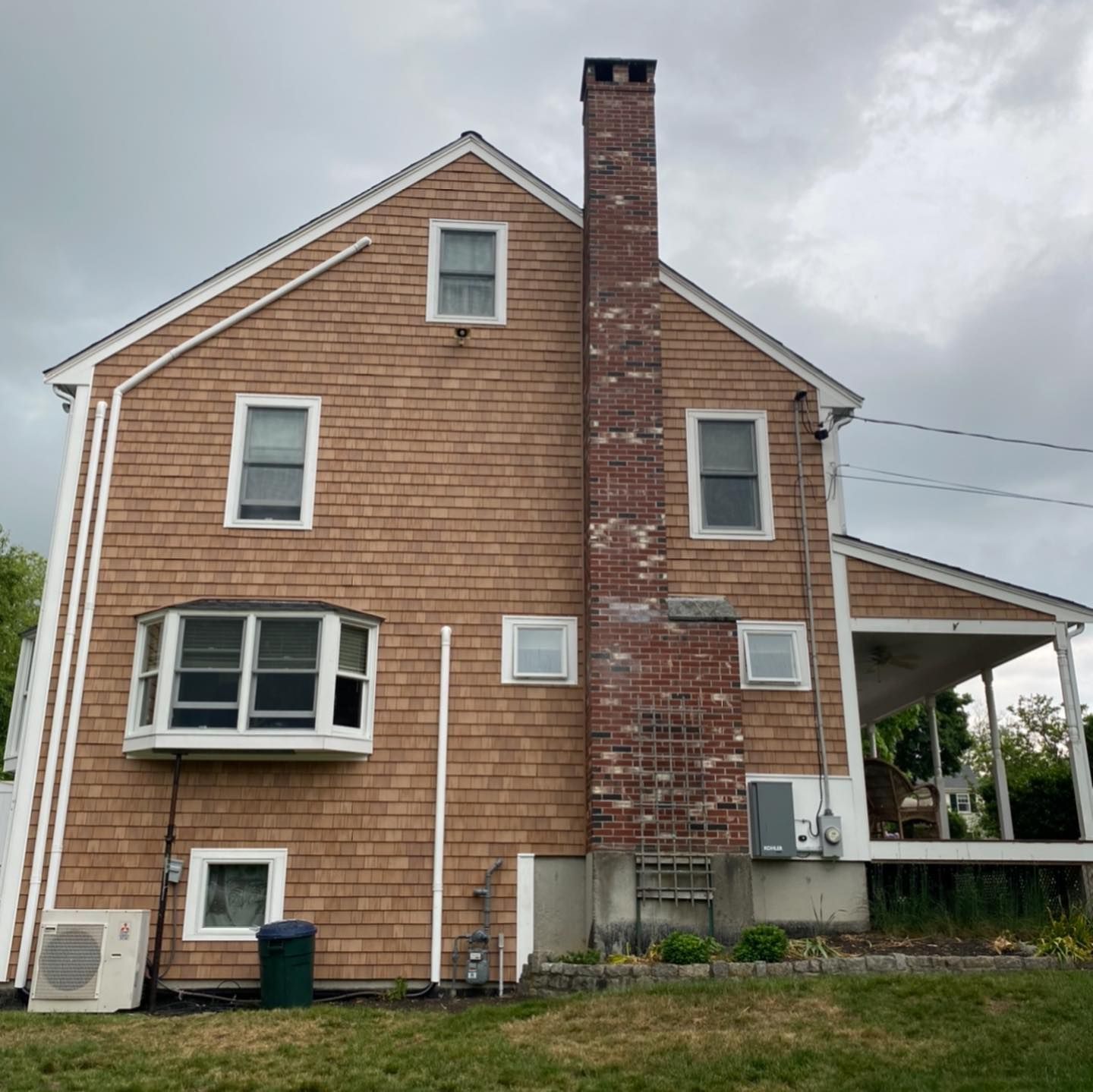A two-story house with light brown wood shingles, a brick chimney, a bay window, and a side porch on a cloudy day.