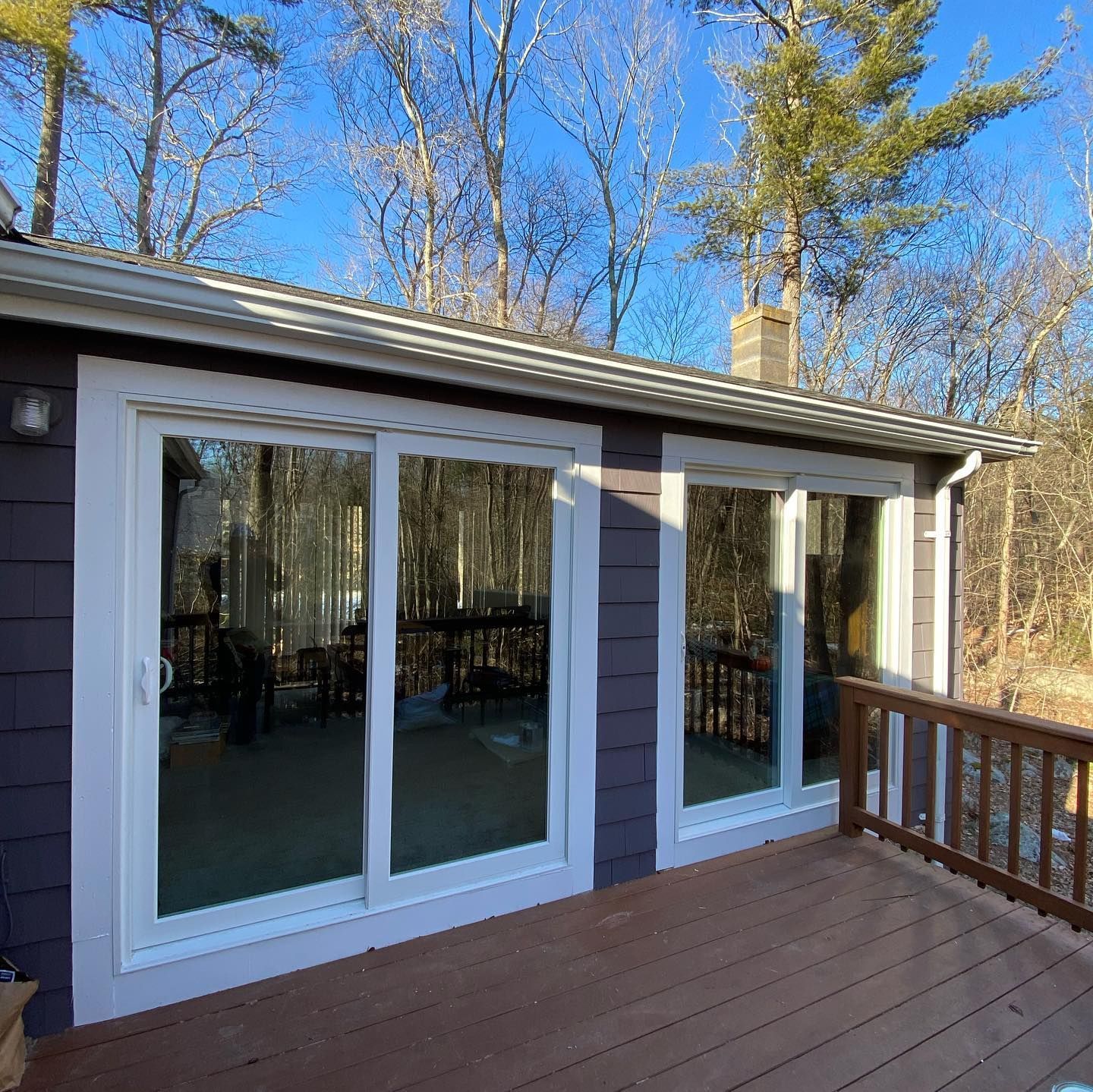 A house exterior with white sliding glass doors, dark wood siding, and a wooden deck, overlooking a forest in sunlight.
